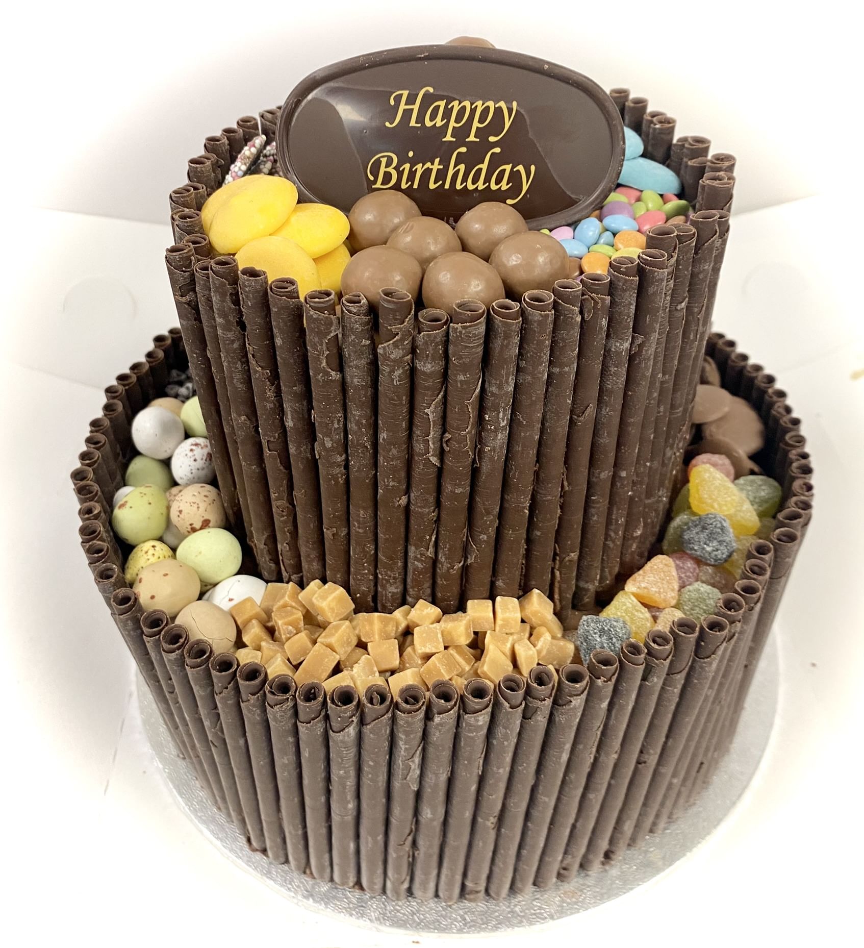 A two-tier chocolate blackout cake decorated with chocolate wafer rolls around the sides. The top tier features a chocolate oval reading 'Happy Birthday,' surrounded by various candies, including yellow jelly beans, malt balls, and colorful sprinkles. The lower tier boasts assorted chocolate candies.